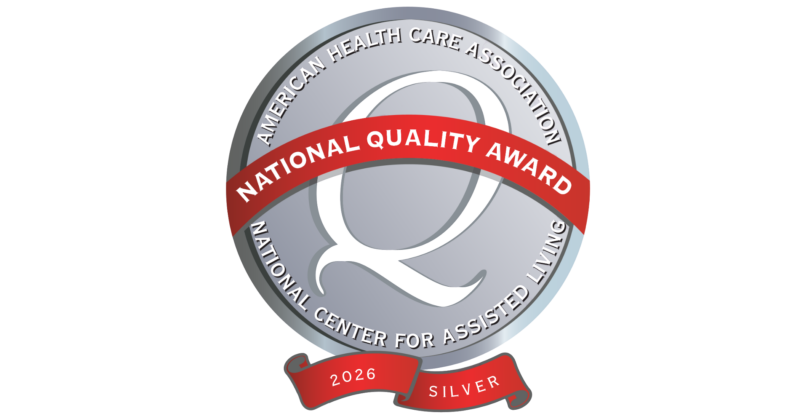 A circular badge with a silver background, featuring a large letter "Q" in the center. Around the "Q" are the words "American Health Care Association" and "National Center for Assisted Living." A red ribbon across the badge reads "National Quality Award," with a smaller red banner at the bottom displaying "2026" and "Silver."